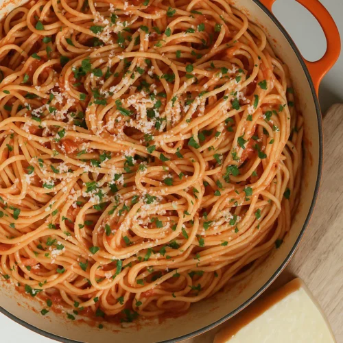 A round pot filled with spaghetti coated in red tomato sauce, garnished with finely chopped green herbs and grated cheese. The pot has an orange handle, and it rests on a wooden cutting board. A wedge of cheese is placed nearby on the board. The background is a marble surface.
