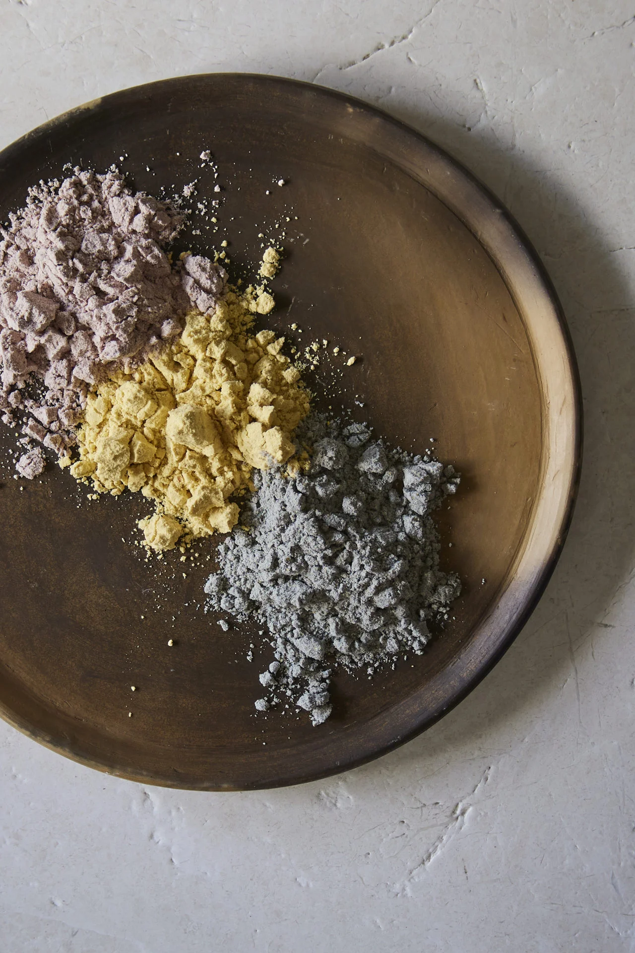 A round wooden plate holds three piles of powder in different colors—pale purple, yellow, and gray—on a light, textured stone surface. The rustic display evokes the artisanal touch found in homemade sopes.