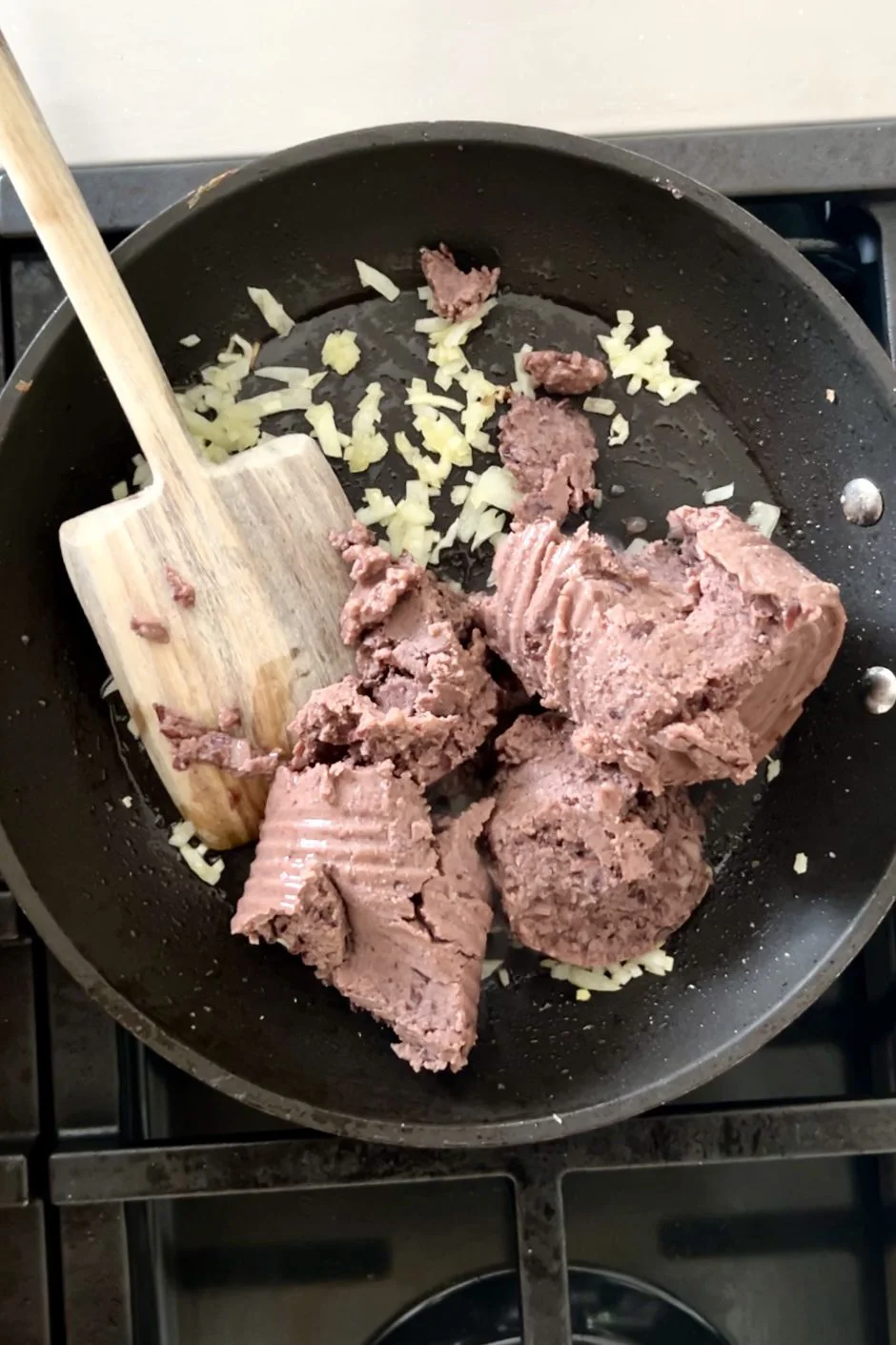 A skillet on a stovetop contains chopped onions being sautéed with several scoops of canned corned beef. A wooden spatula rests in the pan, mixing the ingredients, similar to how you might reheat canned refried beans on the stove.