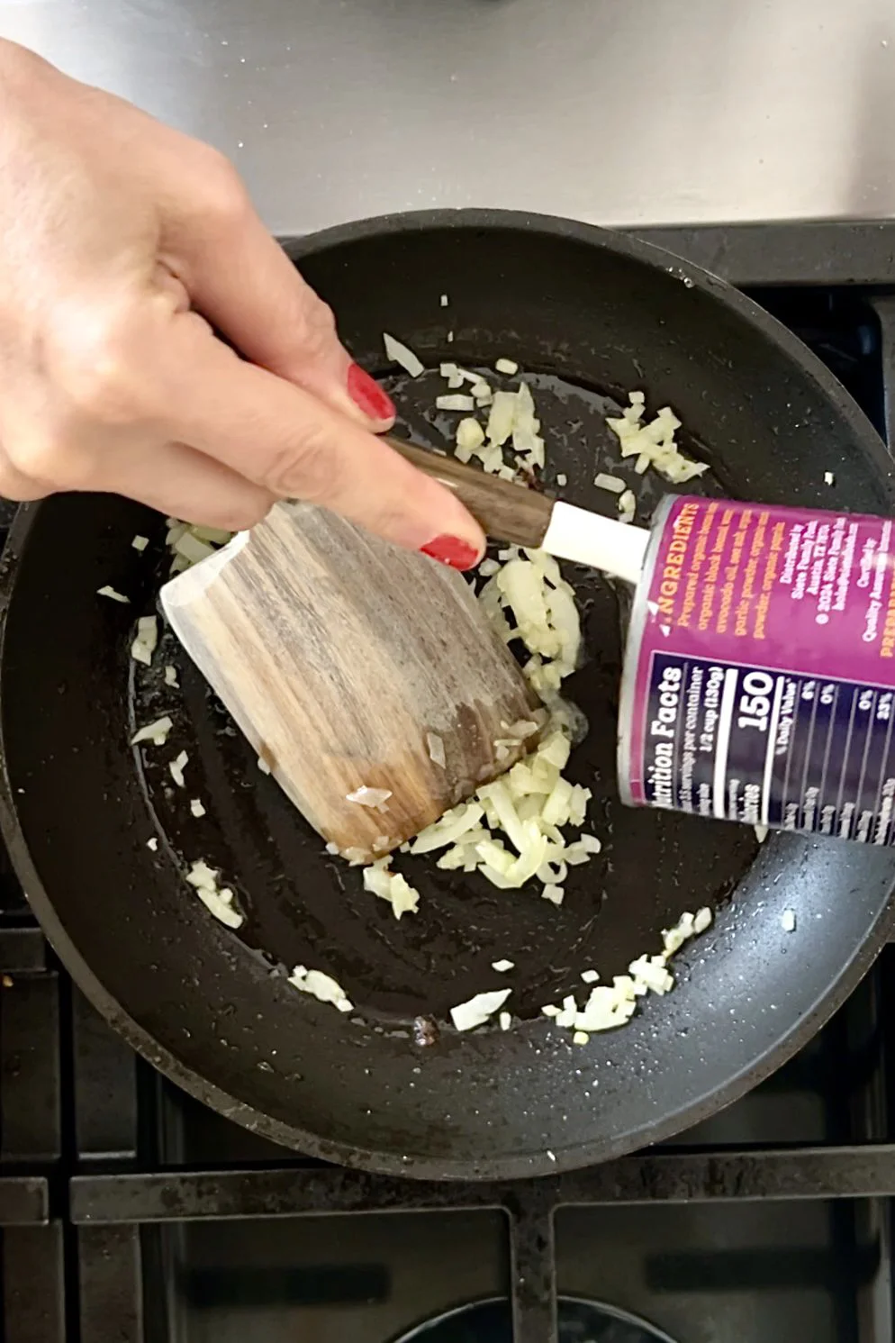 A hand with red nail polish stirs chopped onions in a black frying pan while another pours salt from a purple container—perfect prep for homemade refried beans or before you reheat canned refried beans on the stovetop.