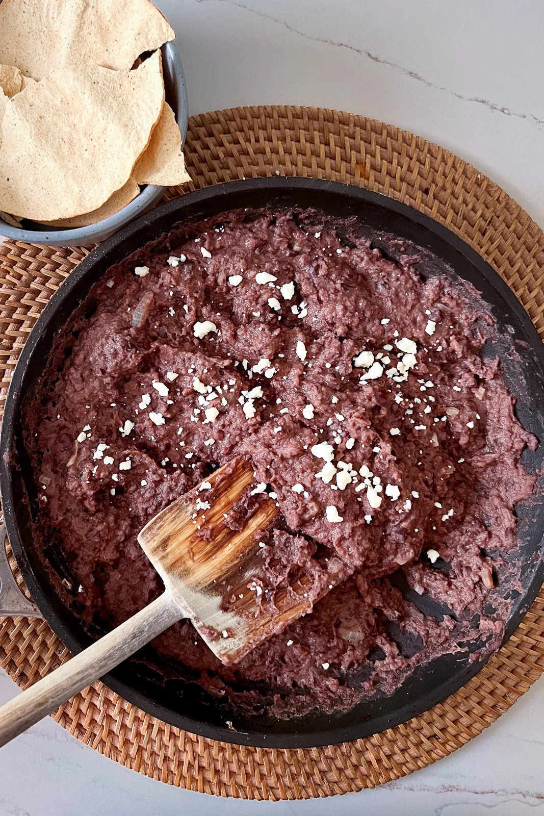 A skillet with creamy homemade refried beans topped with crumbled white cheese sits on a woven placemat. A wooden spoon rests inside the beans. In the top left, a bowl holds crispy tortilla chips on a light-colored table.