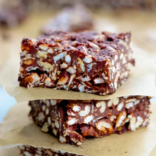 Three vegan cereal chocolate bars stacked on pieces of brown parchment paper. The bars are dense, with visible puffed rice and chunks coated in glossy chocolate, creating a textured look. The blurred background highlights the bars in front.