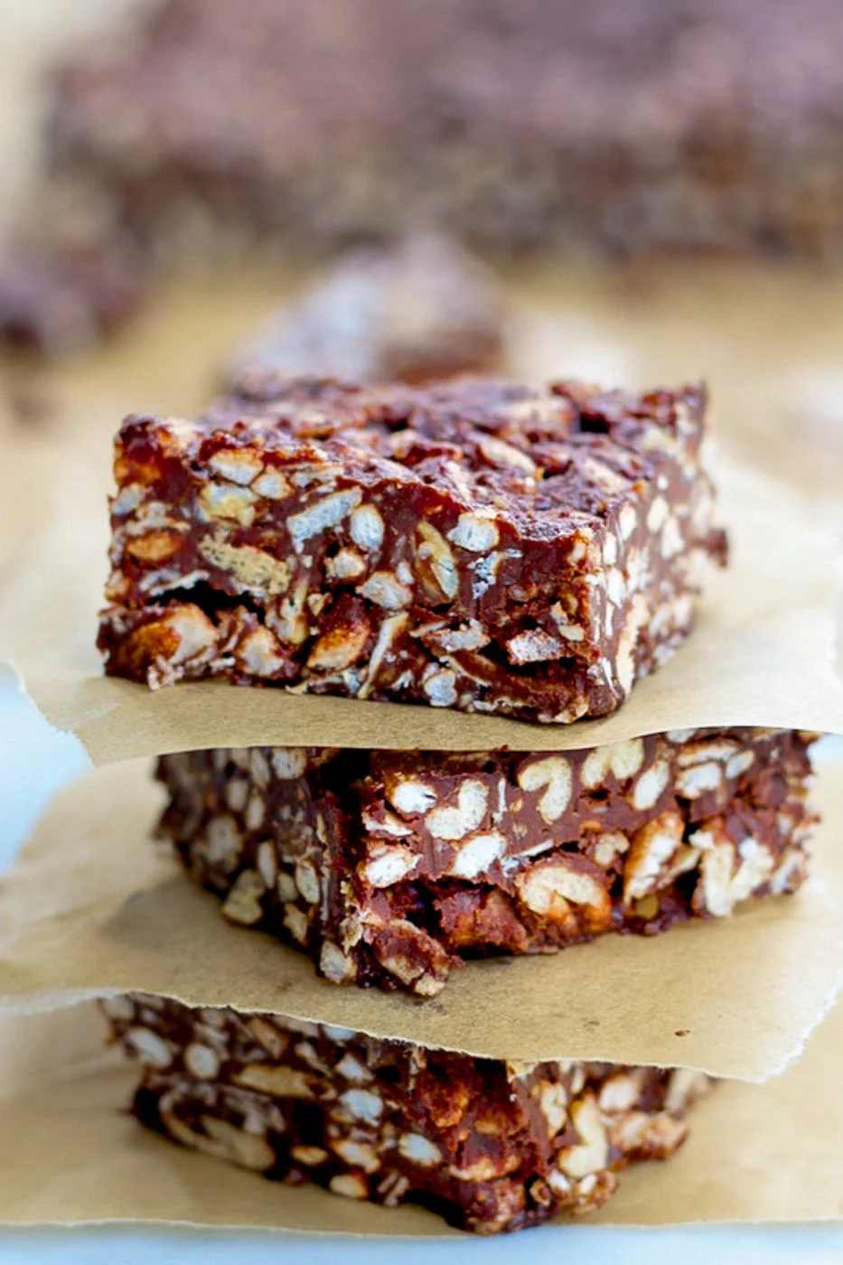 Tres barras veganas de cereal con chocolate apiladas sobre papel pergamino. Las barras son densas, con cereal inflado visible y trozos cubiertos en chocolate brillante, creando una textura visual atractiva.