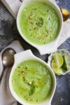Two bowls of creamy avocado and cucumber cold soup are garnished with chopped nuts, a drizzle of oil, and a mint leaf. Nearby rest a vintage spoon, white napkin, and a small dish of lime wedges on the textured dark surface.