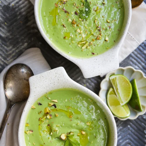 Two bowls of creamy avocado and cucumber cold soup are garnished with chopped nuts, a drizzle of oil, and a mint leaf. Nearby rest a vintage spoon, white napkin, and a small dish of lime wedges on the textured dark surface.