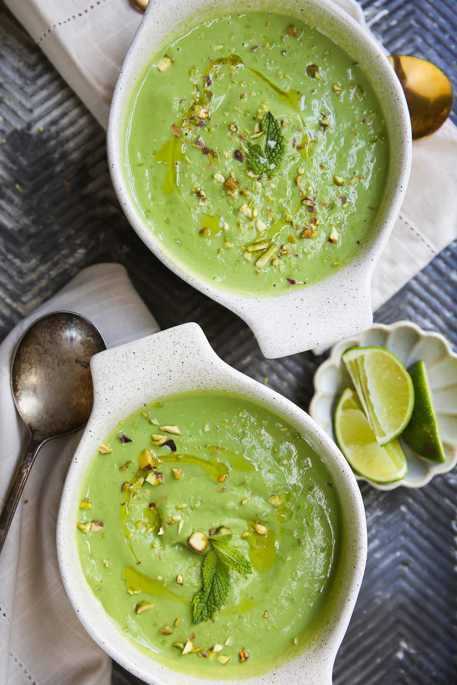 Two bowls of creamy avocado and cucumber cold soup are garnished with chopped nuts, a drizzle of oil, and a mint leaf. Nearby rest a vintage spoon, white napkin, and a small dish of lime wedges on the textured dark surface.