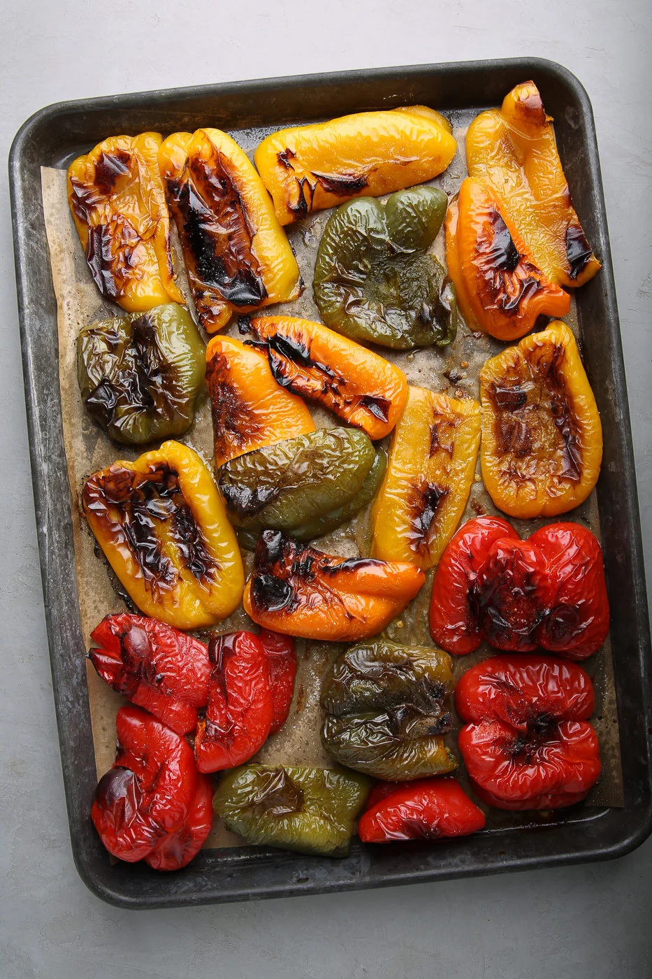A baking tray filled with roasted peppers in red, yellow, and green. The slightly charred and wrinkled halves are browned from the heat—showing exactly how to roast bell peppers. The tray rests on a light gray surface.