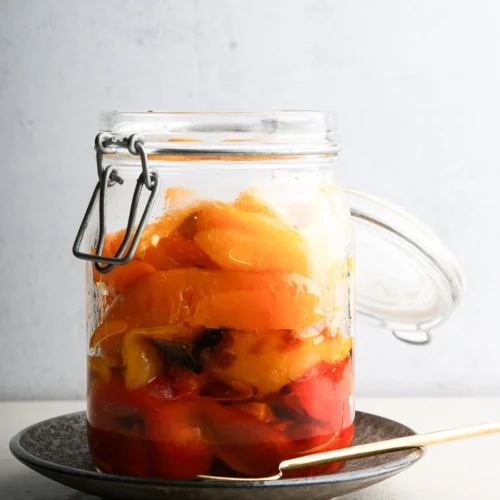 A glass jar filled with colorful roast bell peppers, mostly yellow and red, sits on a dark ceramic plate. The jar's metal clasp lid is open beside it. A fork lies on the plate, all set against a plain, light gray wall.
