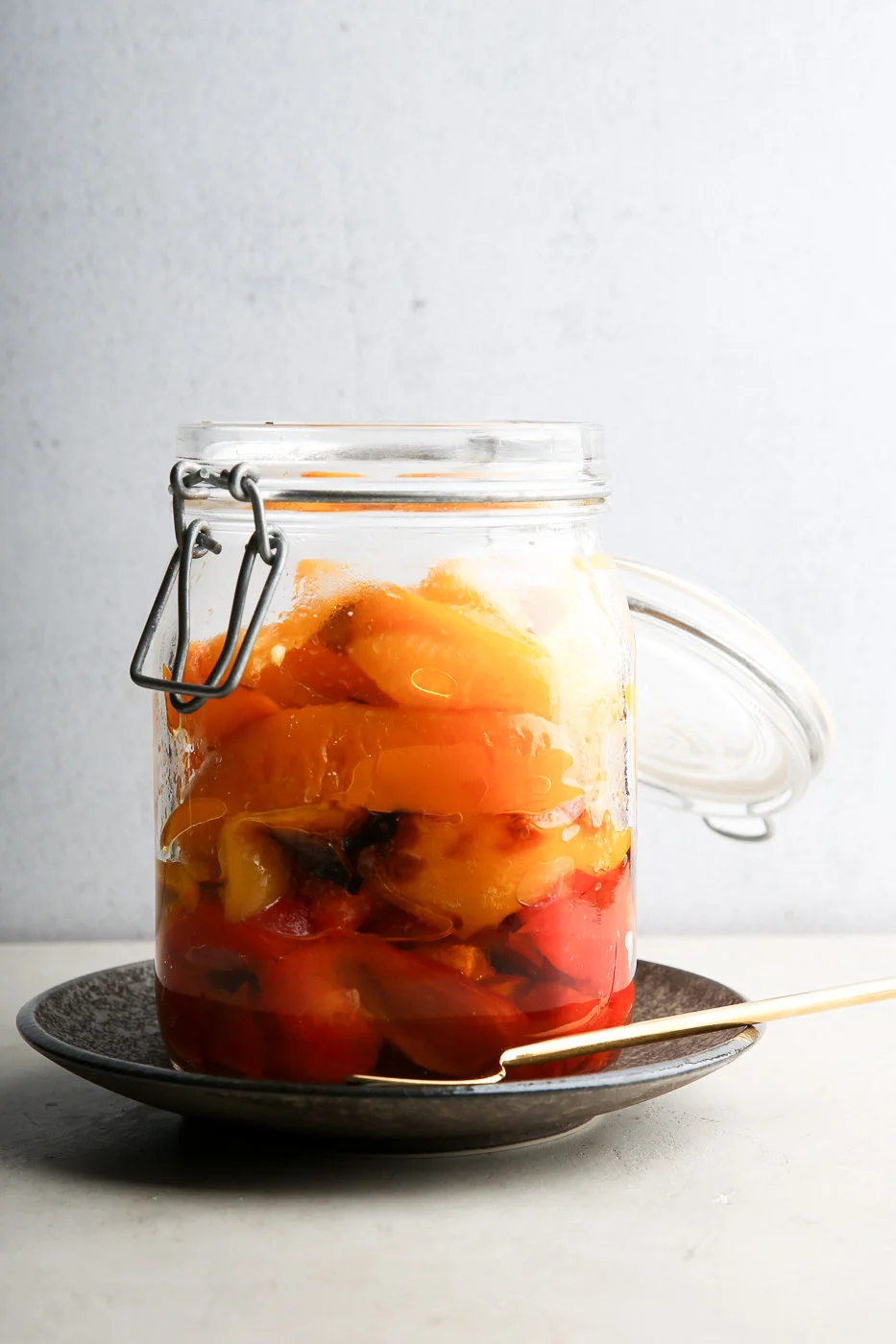 A glass jar filled with colorful roast bell peppers, mostly yellow and red, sits on a dark ceramic plate. The jar's metal clasp lid is open beside it. A fork lies on the plate, all set against a plain, light gray wall.