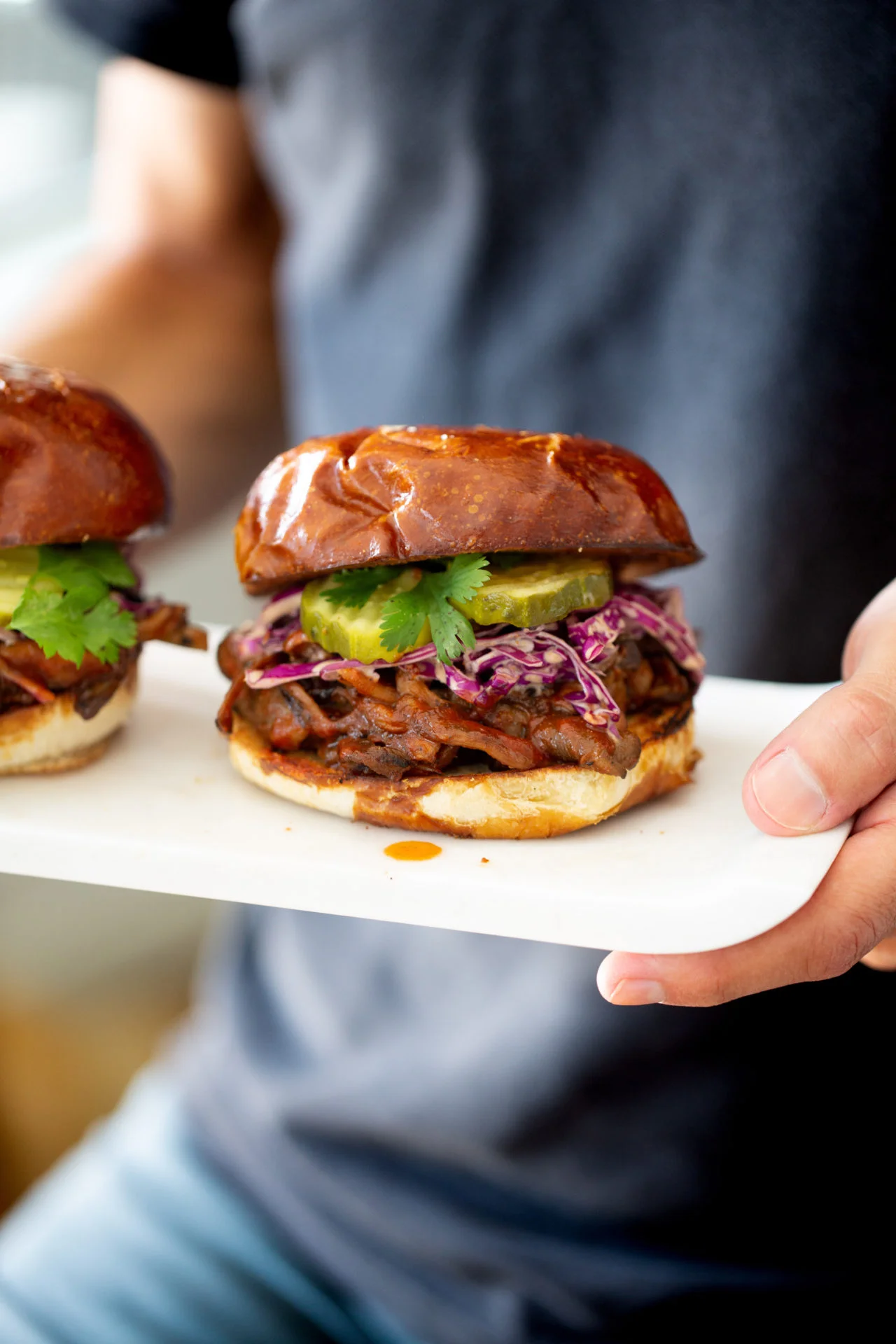 A person holds a white plate with BBQ mushroom sliders on shiny buns, topped with purple slaw, pickles, and cilantro. Another similar sandwich is partially visible. The person wears a dark shirt and blue jeans.