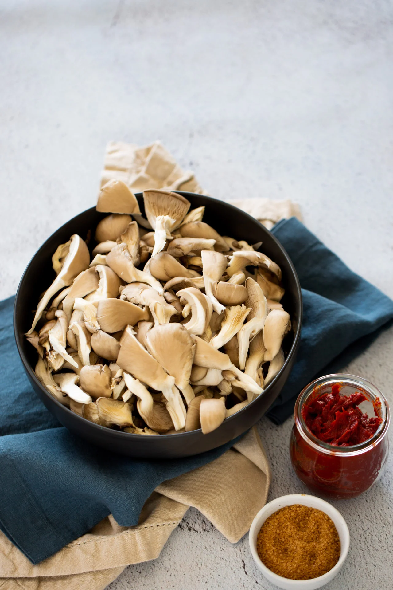 A black bowl filled with chopped oyster mushrooms for BBQ mushroom sliders sits on layered blue and beige cloth napkins. Next to the bowl are a small glass jar of red tomato paste and a white dish containing brown sugar on a light gray surface.