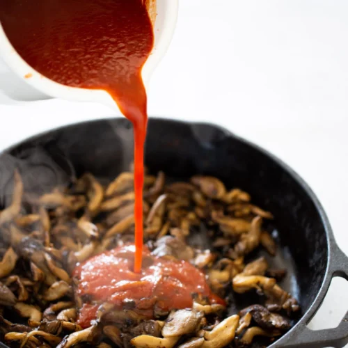 A white cup pours vibrant red homemade Chipotle BBQ sauce into a black cast iron skillet filled with sautéed brown mushrooms, releasing steam. The neutral, light background highlights this vegan cooking process.