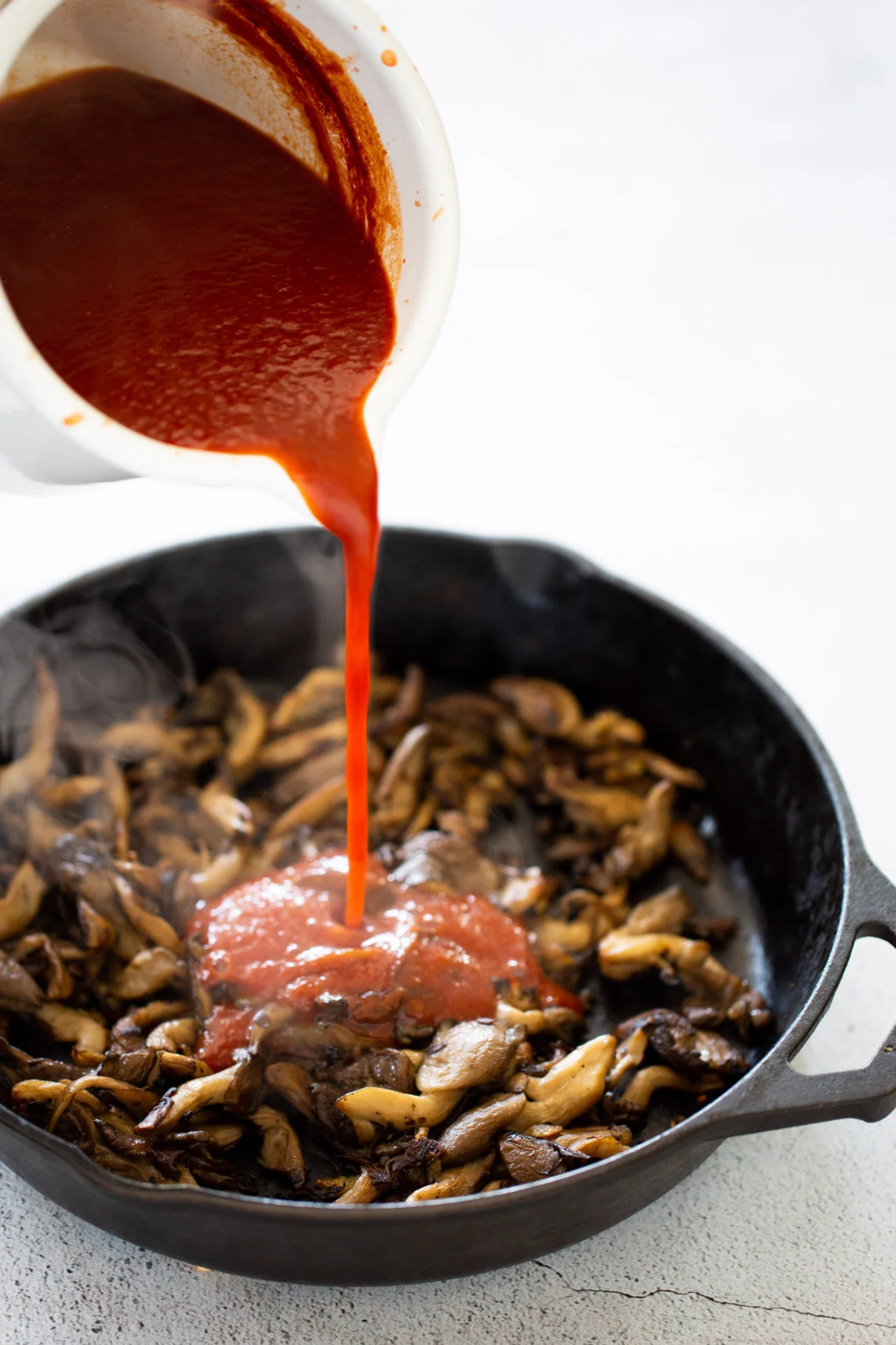 A white cup pours vibrant red homemade Chipotle BBQ sauce into a black cast iron skillet filled with sautéed brown mushrooms, releasing steam. The neutral, light background highlights this vegan cooking process.