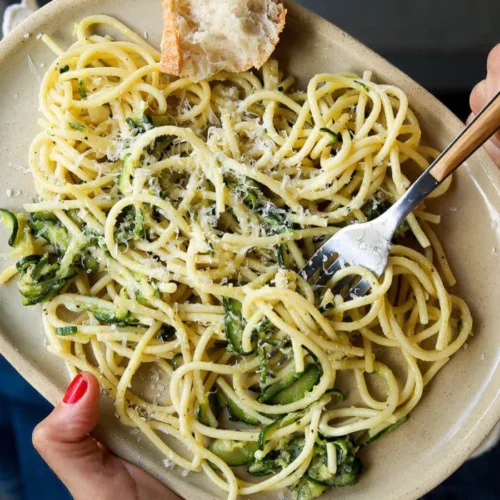 A person holds an oval plate of Zucchini Parmesan Pasta—a quick & easy summer recipe with green veggies and grated cheese. A fork twirls pasta, a rustic bread sits on the side, and the person wears red nail polish, blue jeans, and a white sweater.