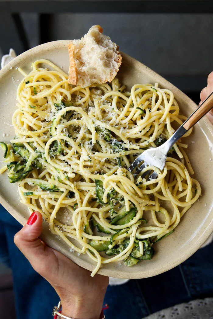 A person holds an oval plate of Zucchini Parmesan Pasta—a quick & easy summer recipe with green veggies and grated cheese. A fork twirls pasta, a rustic bread sits on the side, and the person wears red nail polish, blue jeans, and a white sweater.