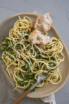 An oval plate holds a serving of Quick and Easy Zucchini Parmesan Pasta, topped with grated cheese and two pieces of rustic bread. A fork sits beside the plate, which rests on a light-colored cloth—perfect as a summer recipe.