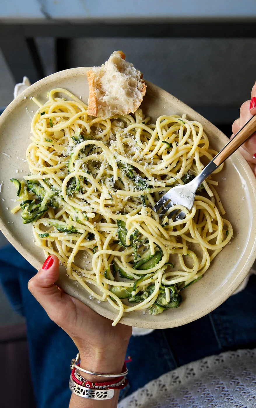 A person with red nail polish holds a beige oval plate of Zucchini Parmesan Pasta—a quick & easy summer recipe—twirling a fork in the cheesy spaghetti while crusty bread rests on the edge. They wear a white lace top, jeans, and layered bracelets.