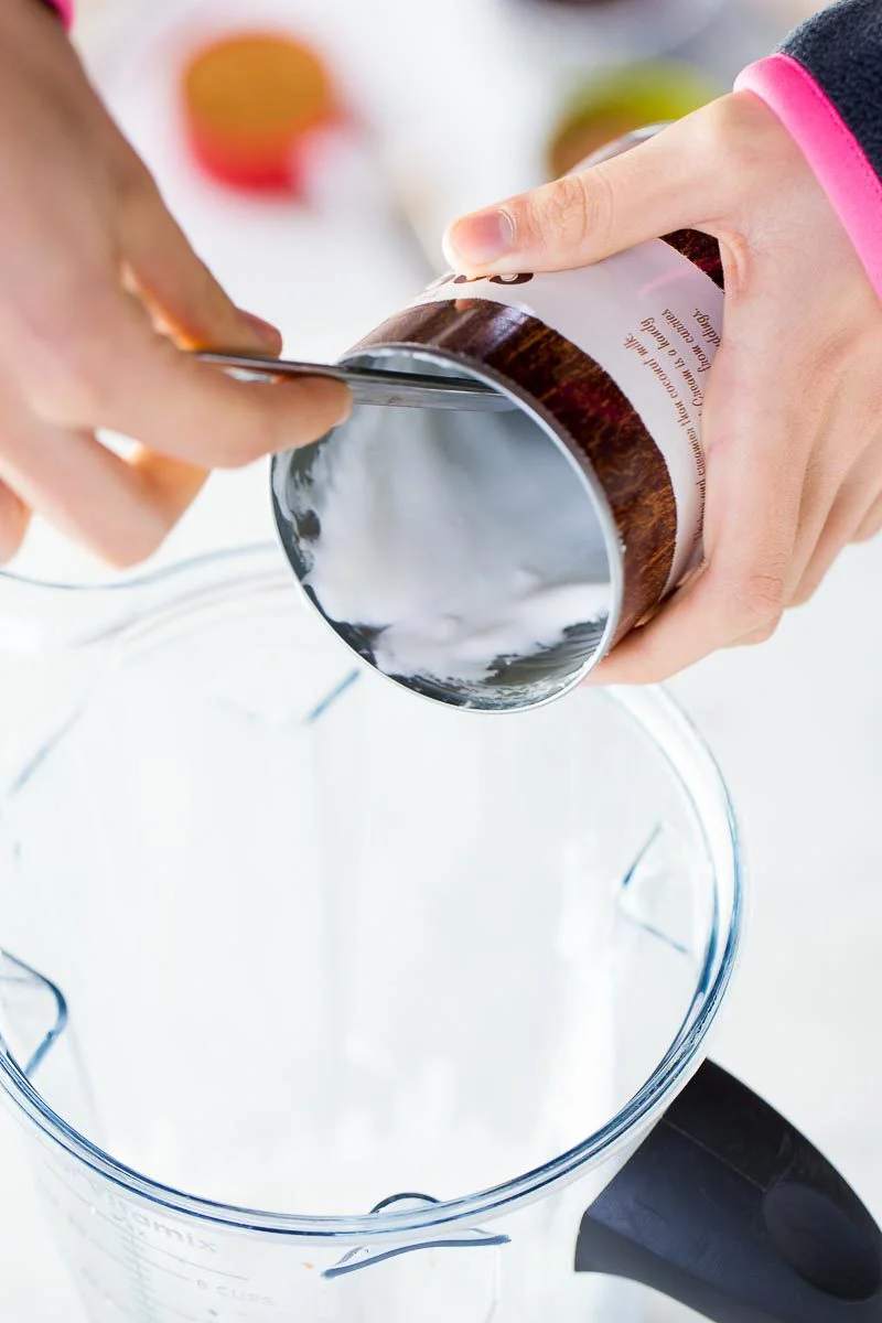 Una persona vierte leche de coco espesa en una licuadora, preparando la mezcla para un helado vegano de chocolate. Sus manos, con una blusa rosa, están enfocadas sobre un fondo claro y desenfocado.