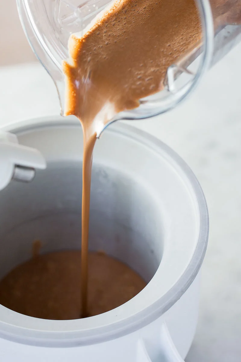 Mezcla líquida de helado vegano de chocolate siendo vertida desde una licuadora a un tazón blanco de máquina para helado, lista para batirse.