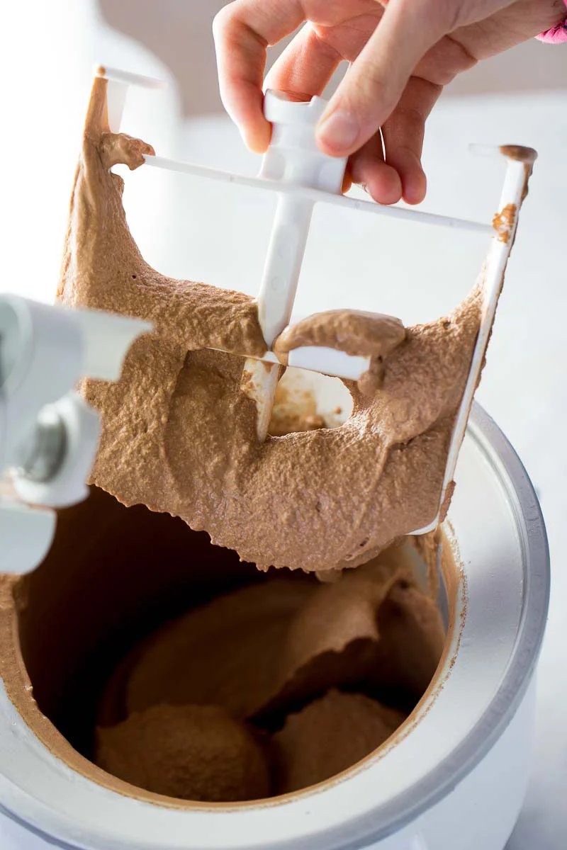 Una mano sostiene la pala mezcladora cubierta de helado vegano de chocolate recién batido, sobre el tazón de la máquina de helado.