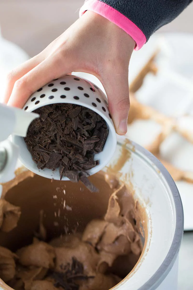 Una mano vierte una taza de chocolate oscuro picado dentro de la máquina para helado con helado vegano de chocolate. La taza blanca con puntos negros da un toque divertido.