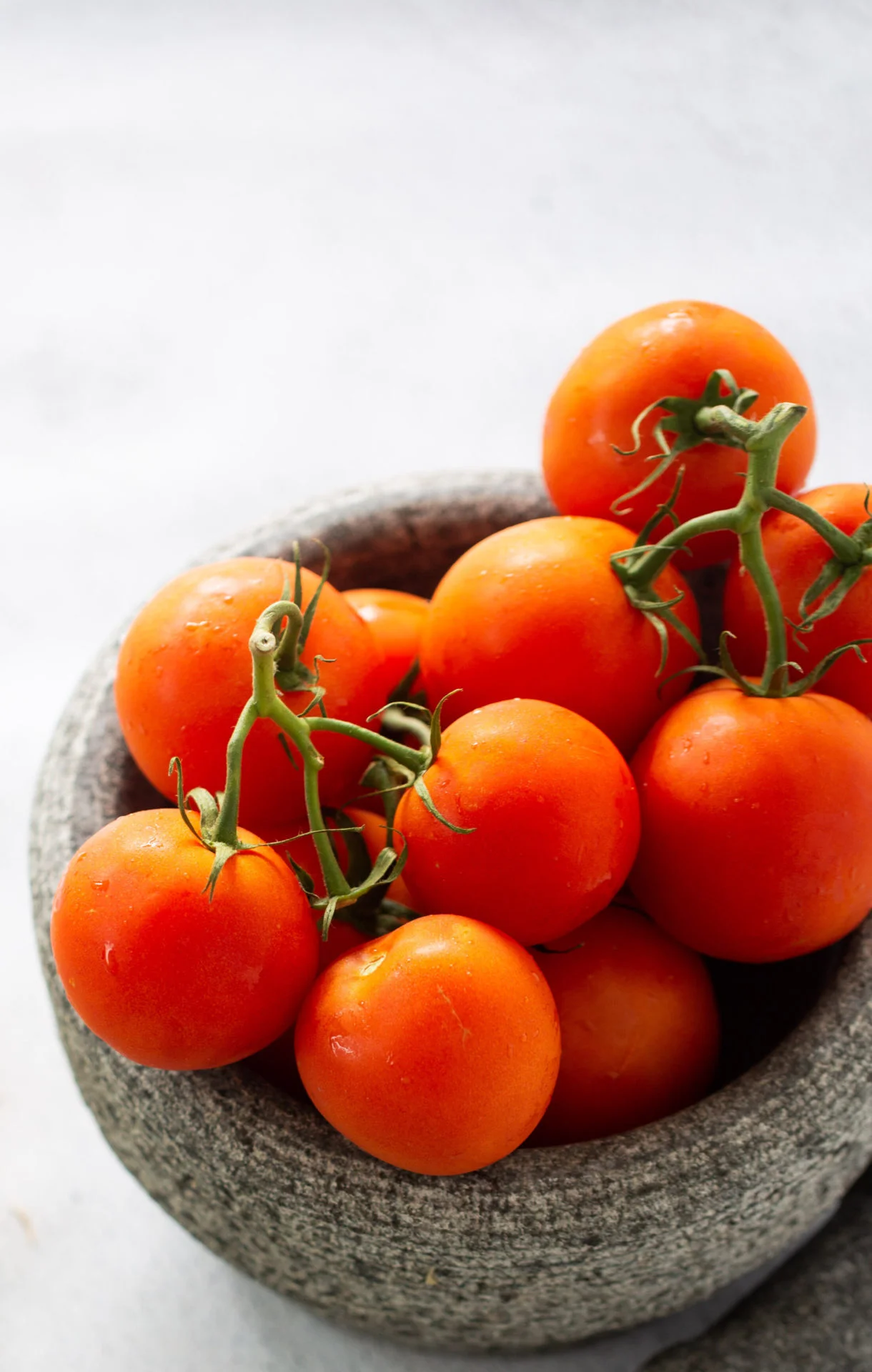 A gray stone bowl filled with ripe, red tomatoes—perfect for a homemade tomato sauce recipe—some still attached to green vines. The bowl sits on a light, textured surface, and the tomatoes appear fresh with a slight sheen of moisture.