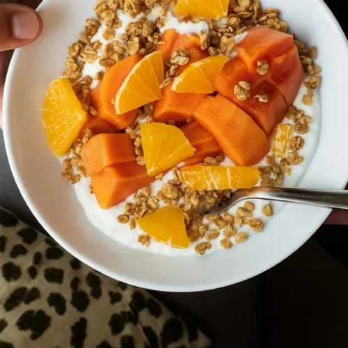 A hand holds a papaya breakfast bowl filled with yogurt, granola, orange slices, and chunks of papaya above a leopard print cushion. A spoon rests inside this vibrant Mexican breakfast.