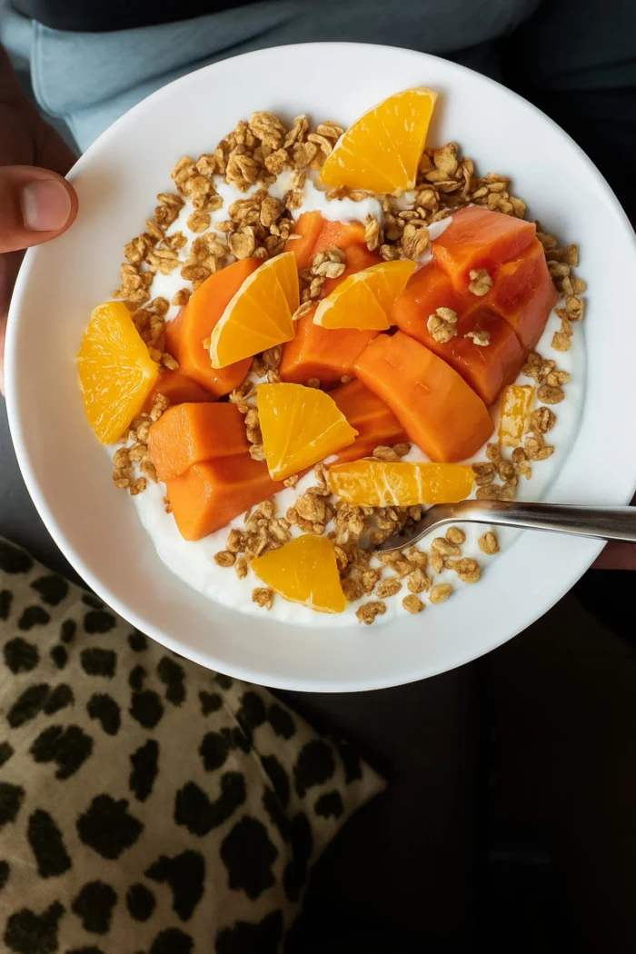 A hand holds a papaya breakfast bowl filled with yogurt, granola, orange slices, and chunks of papaya above a leopard print cushion. A spoon rests inside this vibrant Mexican breakfast.