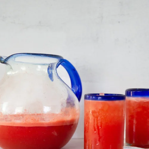 A glass pitcher with a blue handle, filled halfway with bright red Mexican Watermelon Water, sits on a white surface. Beside it are two tall glasses of refreshing Watermelon Agua Fresca on folded white napkins, against a light, neutral background.