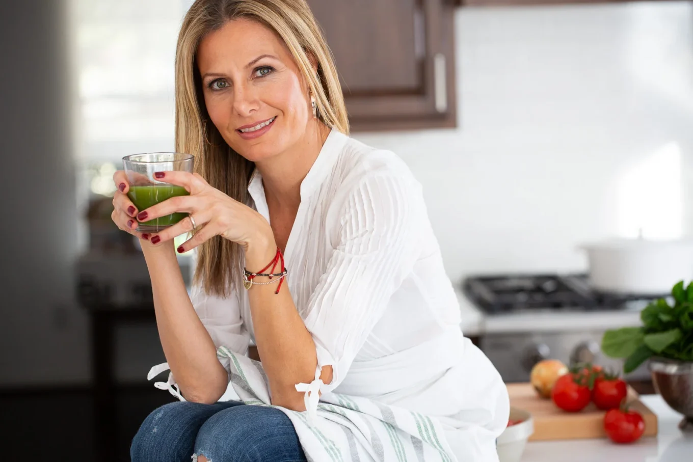A woman with blonde hair and a white blouse sits in a kitchen, smiling and holding a glass of green juice. Fresh vegetables, including tomatoes and spinach, are on the counter beside her. The kitchen has brown cabinets and a white stove in the background.