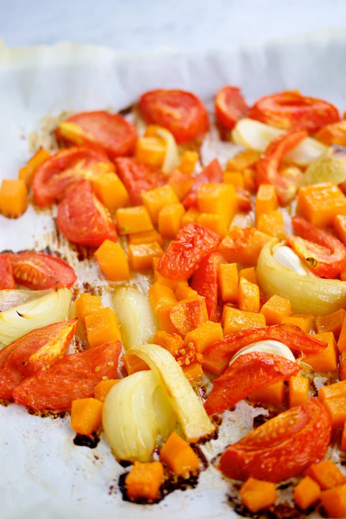 Chunks of orange butternut squash and red tomatoes are roasted with yellow onion wedges on a parchment-lined baking sheet, creating the perfect base for a flavorful butternut squash pasta sauce. The veggies are soft, caramelized, and lightly browned on the edges.
