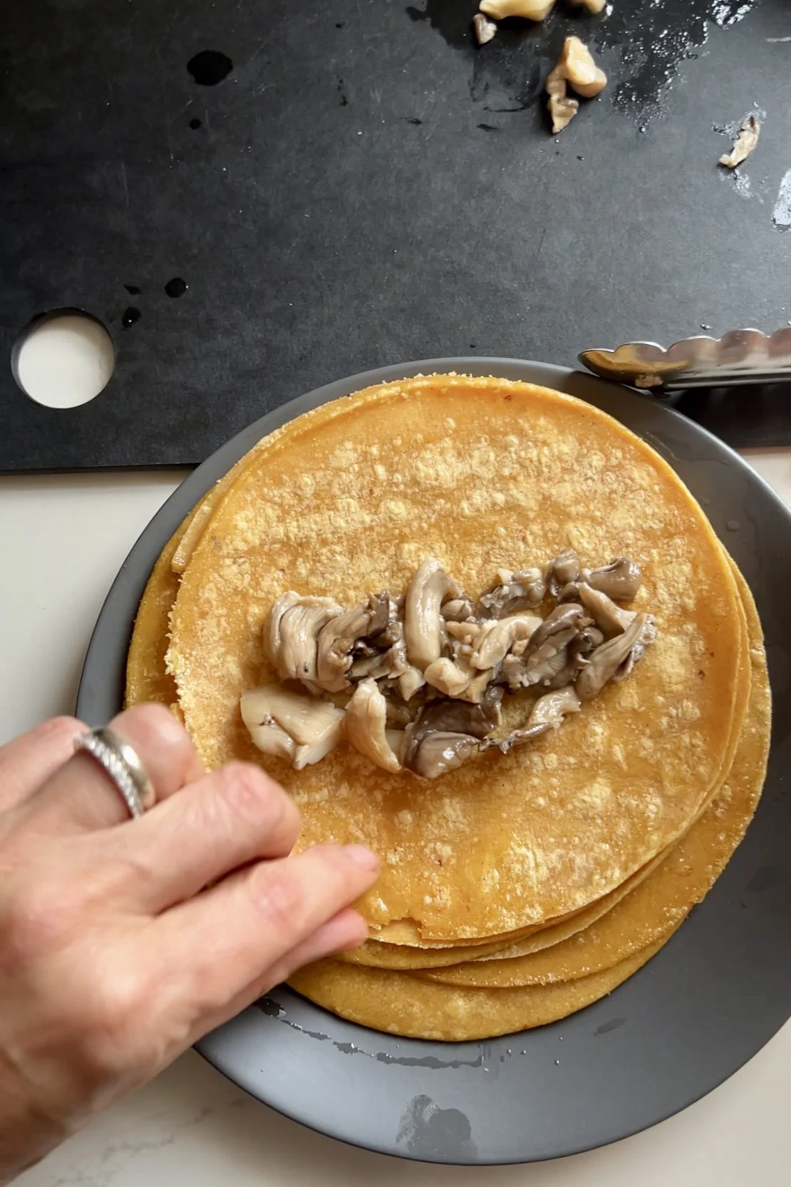 A hand prepares to fold a stack of yellow corn tortillas with sautéed oyster mushrooms on top. The plate rests on a white surface, with metal tongs and a black cutting board with food scraps visible in the background.
