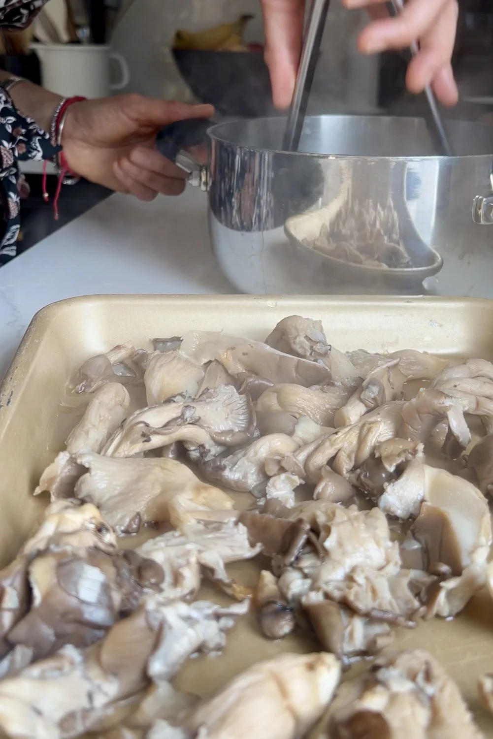 A beige tray filled with chopped oyster mushrooms sits in the foreground. In the background, a person in patterned clothing holds a large silver pot over a stove, with steam rising—hinting at enchiladas verdes or another dish in progress.