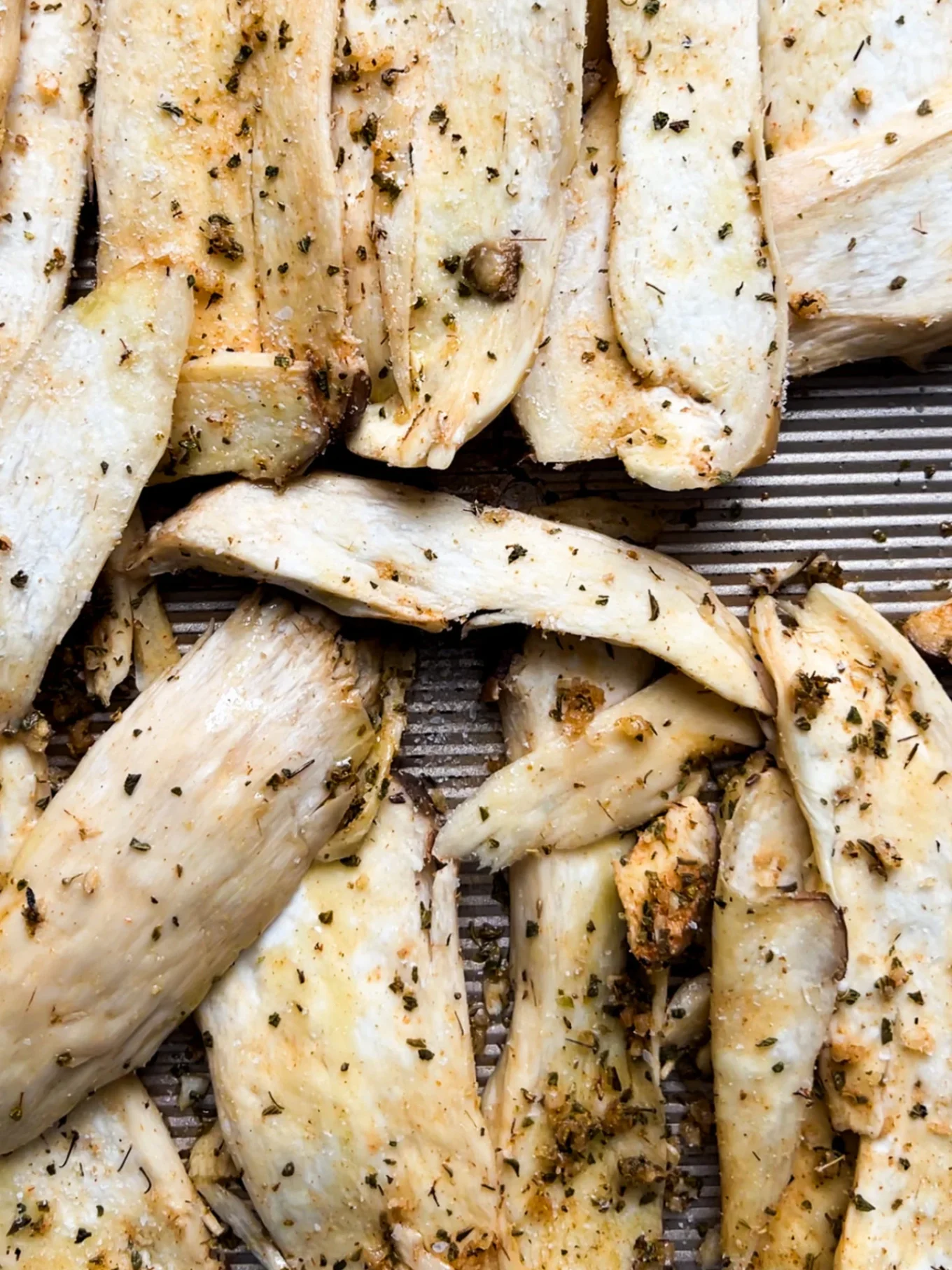 Sliced, roasted king oyster mushrooms are arranged on a ridged baking tray. Perfect for a vegetarian gyro, these golden brown mushrooms are seasoned with herbs and spices, giving them a flavorful appearance with irregular, crispy edges.