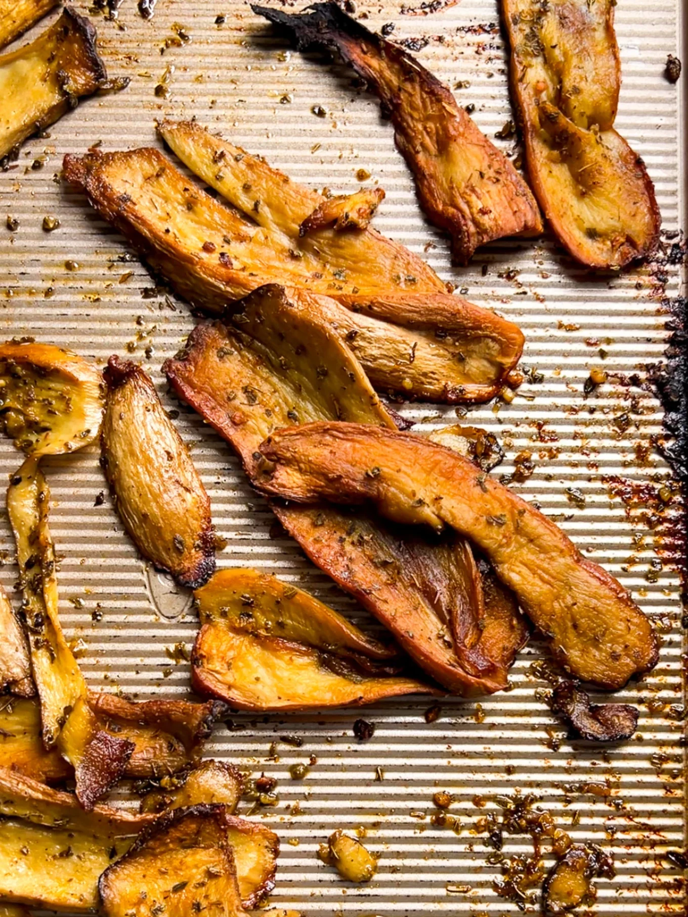 Golden-brown roasted root vegetable slices and King Oyster Mushrooms, seasoned with herbs and spices, are scattered on a ridged baking sheet. The vegetables are caramelized, with some edges slightly crisp and flecked with visible seasoning.