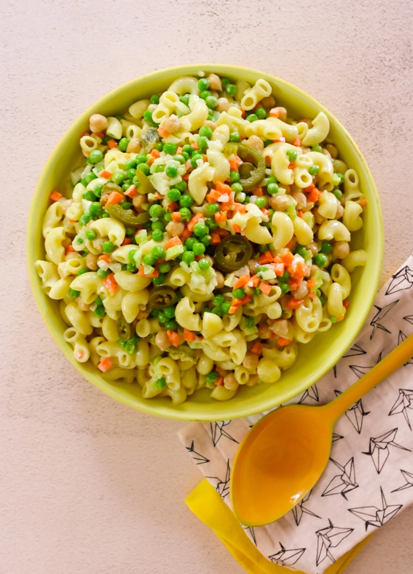 A bowl of Mexican pasta salad with green peas, diced carrots, corn, chickpeas, and sliced olives sits on a white surface. A yellow spoon and a patterned napkin are beside the bowl. The colorful salad looks freshly prepared.