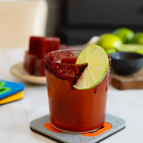 A glass rimmed with chili powder, filled with a Magic Michelada, is garnished with a lime slice. The drink sits on a coaster atop a light surface, with limes and Michelada ice cubes on a plate in the background.