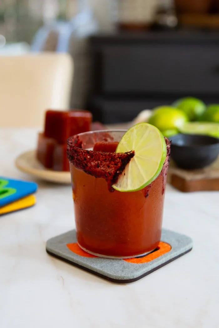 A glass rimmed with chili powder, filled with a Magic Michelada, is garnished with a lime slice. The drink sits on a coaster atop a light surface, with limes and Michelada ice cubes on a plate in the background.