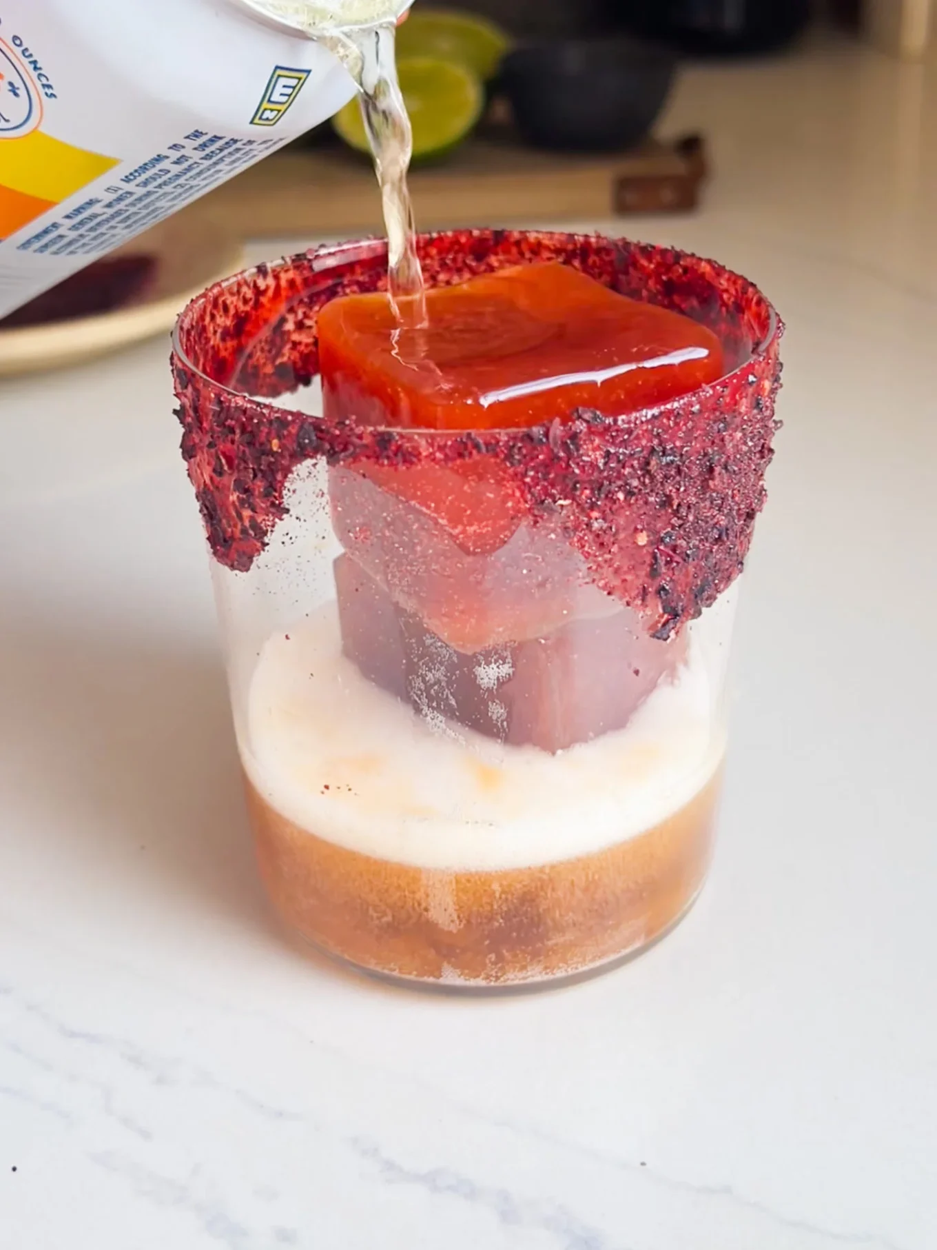 A close-up of a glass rimmed with deep red chili powder and a large Michelada ice cube. A carbonated drink is poured in, creating foam. The scene, perfect for the best Michelada, sits on a white countertop with blurred limes and a dark bowl behind.