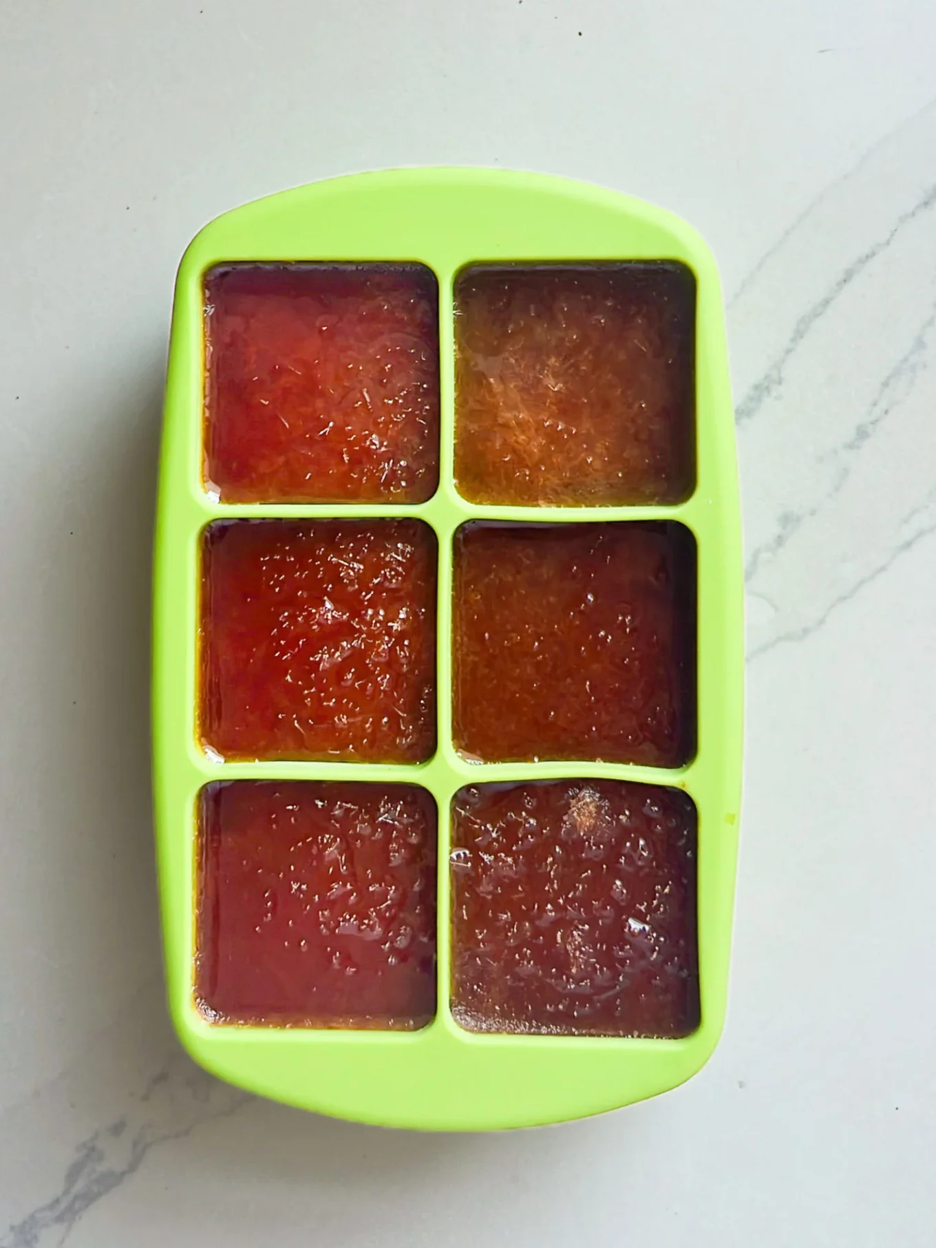 A green silicone ice cube tray filled with six sections of reddish-brown Michelada ice cubes sits on a light, slightly marbled countertop—each section perfectly portioned for the best way to make a Michelada.