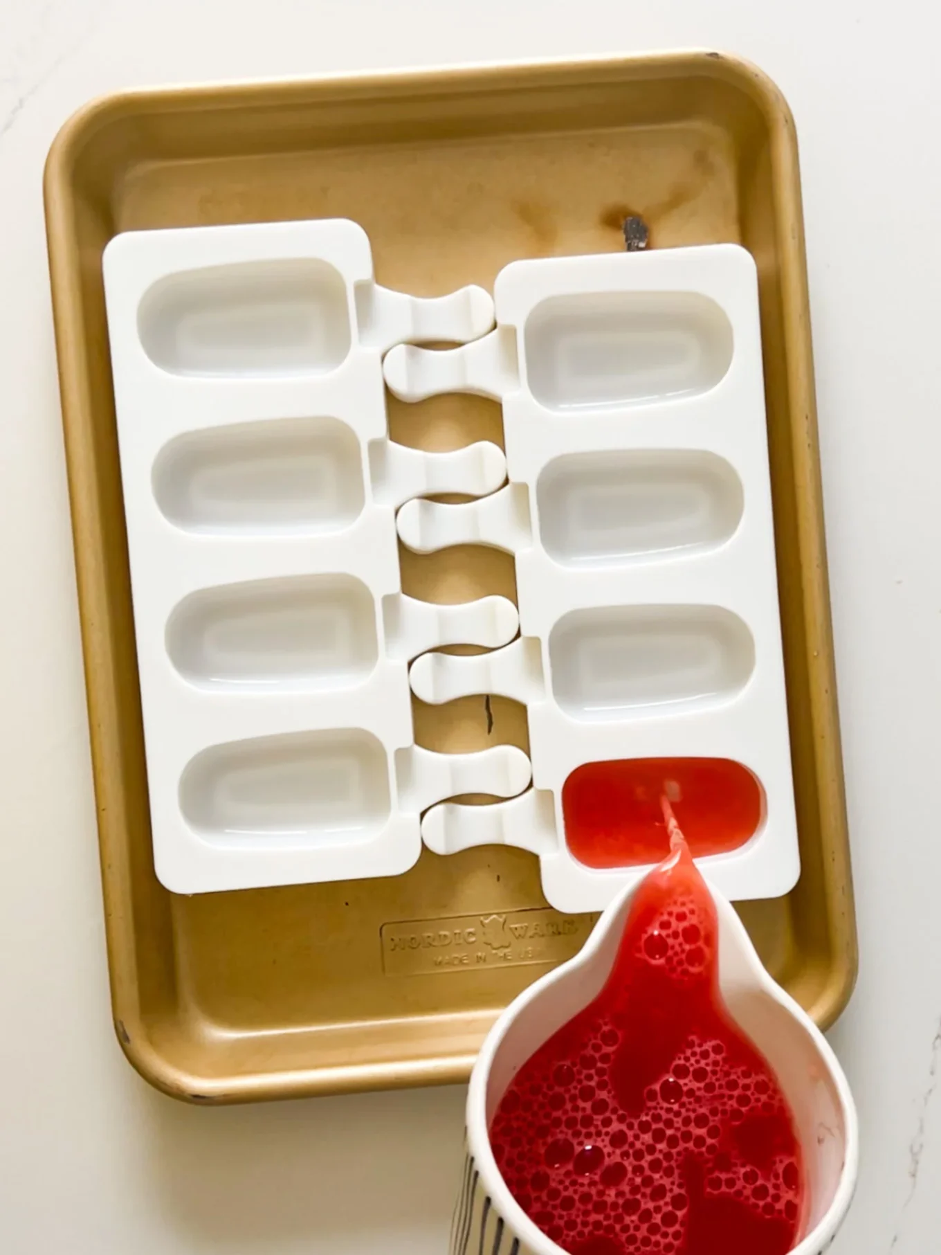 A white silicone popsicle mold with six slots sits on a gold baking sheet. A striped measuring cup pours bright red liquid—perfect for making Watermelon Paletas—into one mold, while the others remain empty on a light-colored surface.