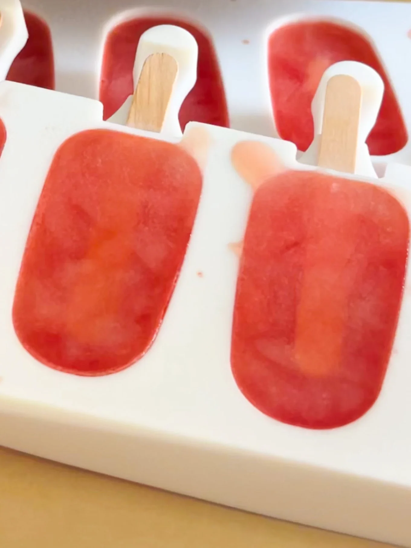 A close-up of a white popsicle mold holding bright red and orange Watermelon Paletas on wooden sticks. Some popsicle liquid is slightly spilled on the mold, and the popsicles appear partially frozen. The background is blurred.