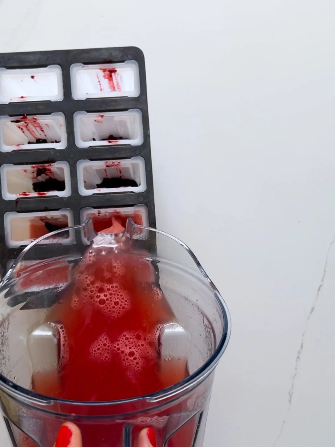 A close-up shows a blender jar with red liquid, possibly Hibiscus Chamoy, being poured into a gray ice cube tray, which already contains some red-stained sections—evoking the making of Watermelon Paletas on a marbled white background.