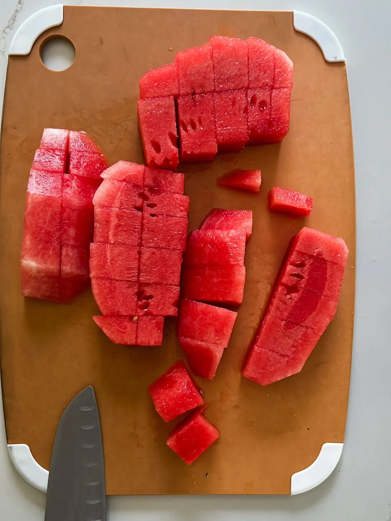 Chunks of bright red watermelon, some with rind removed, are neatly cut and arranged on a brown cutting board with white corners—perfect for making refreshing Watermelon Paletas. A chef’s knife rests in the lower left corner beside the pieces.