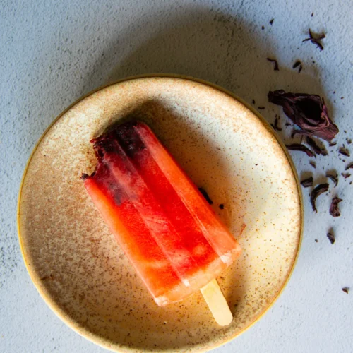 A partially eaten red popsicle with a wooden stick rests on a speckled beige plate. Dried purple flower petals are scattered on the light gray textured surface beside the plate. The scene is bright and minimalistic.