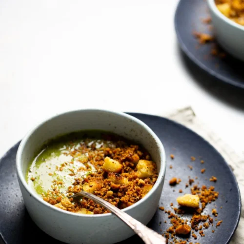 A bowl of creamy green soup inspired by Mexican Soup Recipes is topped with golden croutons and brown crispy crumbs on a black plate. A spoon rests in the bowl, while another bowl is partly visible in the background on a white surface.
