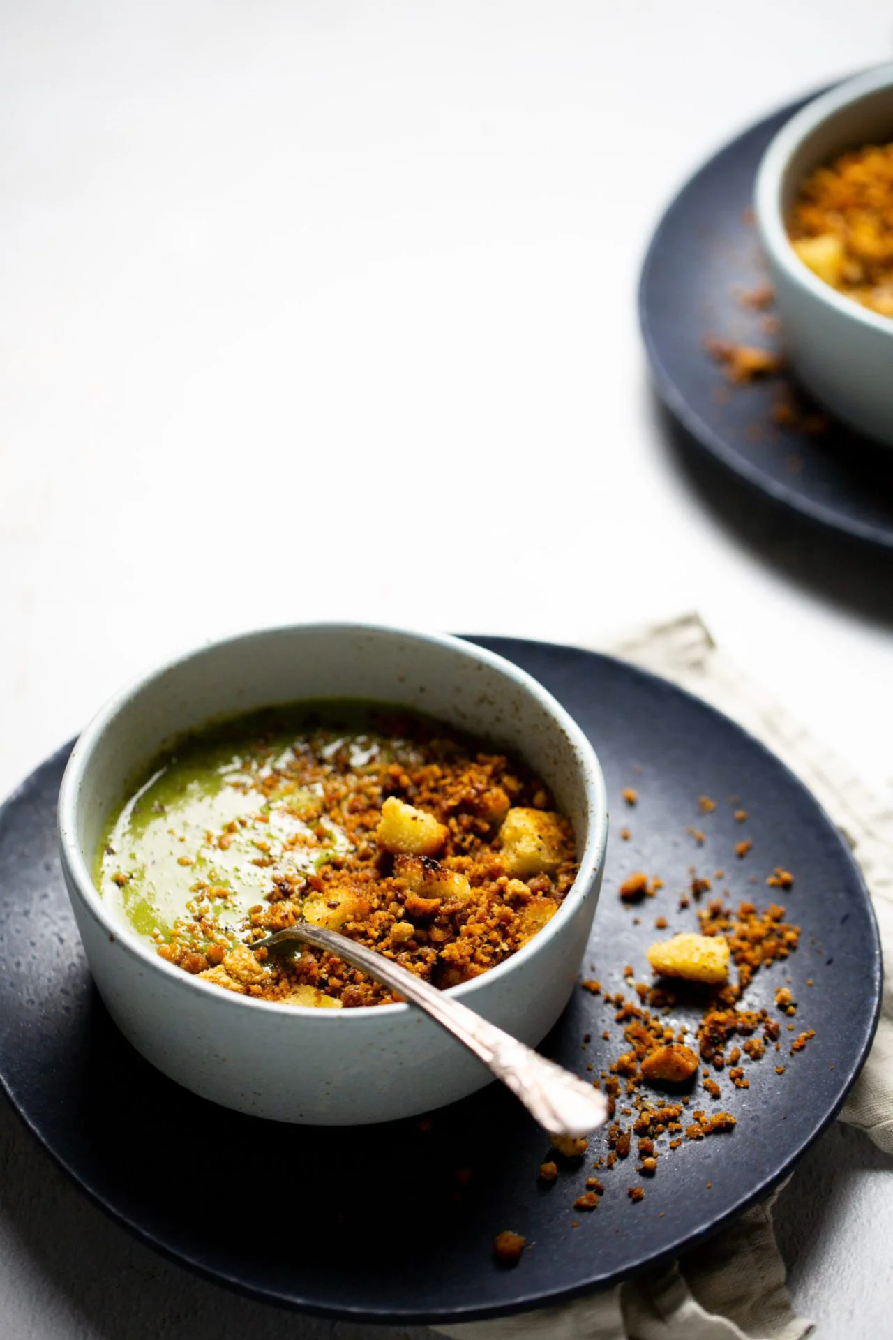 A bowl of creamy green soup inspired by Mexican Soup Recipes is topped with golden croutons and brown crispy crumbs on a black plate. A spoon rests in the bowl, while another bowl is partly visible in the background on a white surface.
