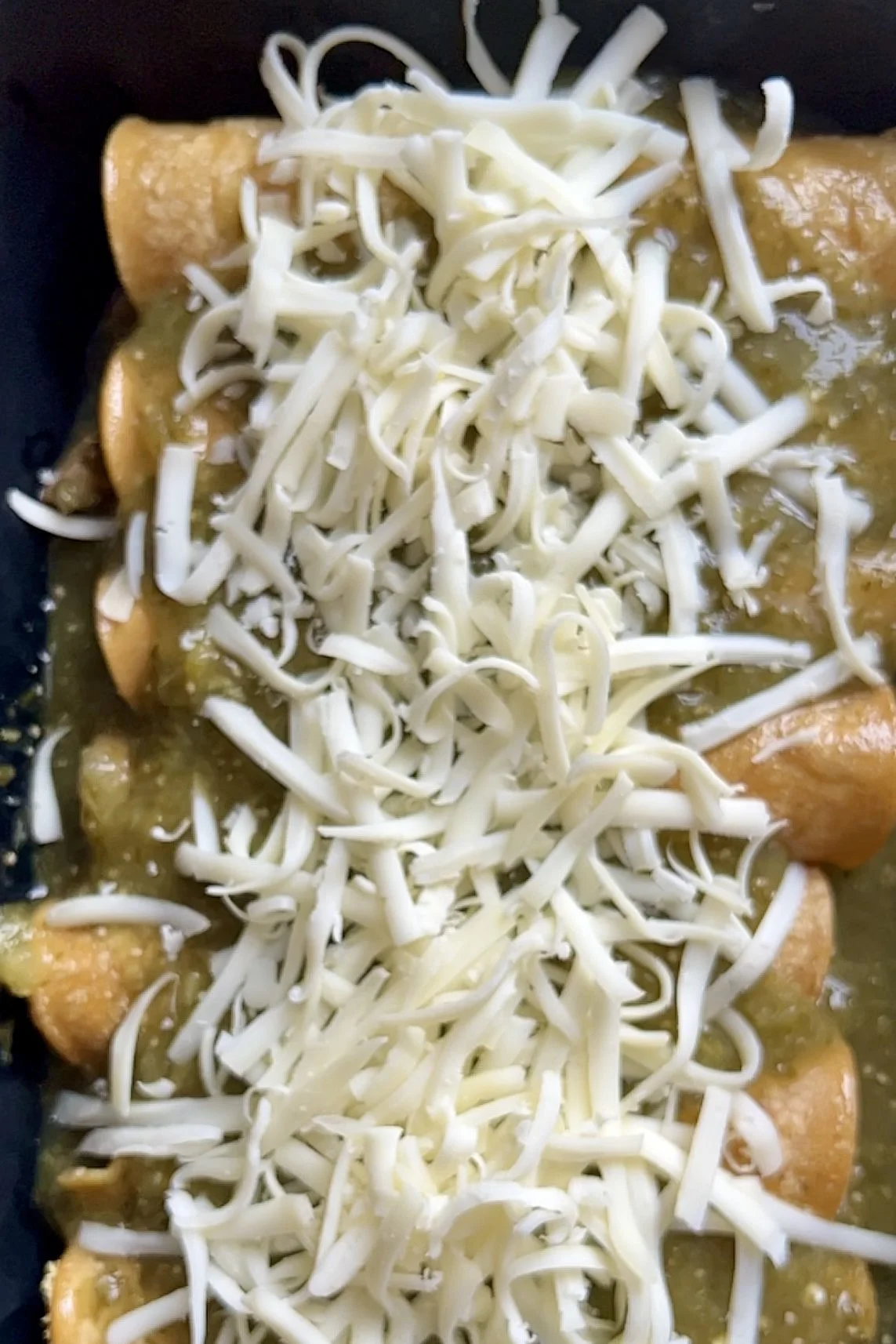 A close-up of enchiladas verdes in a baking dish, covered with shredded white cheese and topped with green salsa. The tortillas are rolled and lined up, with the cheese generously spread across the center.