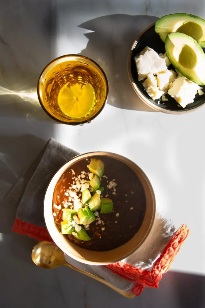 A bowl of Caldo de Frijol topped with diced avocado and cheese rests on a folded napkin beside a gold spoon. Nearby, a glass of amber drink and a small bowl with avocado slices and crumbled cheese sit on a white marble surface in bright sunlight.