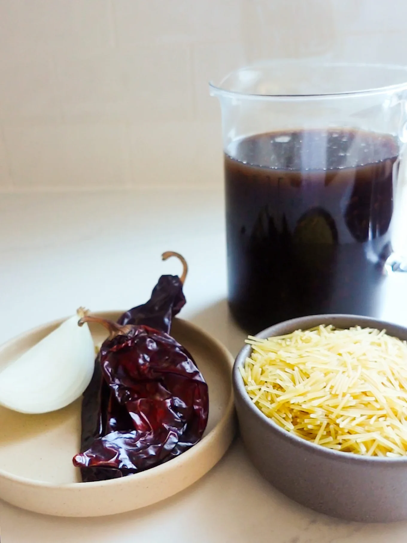 A clear pitcher of dark brown broth sits behind a small beige plate with two dried red chiles and a quarter white onion, next to a bowl filled with uncooked thin yellow noodles—classic ingredients for Mexican soup like Sopa de Fideo.
